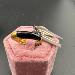 May include: A gold and black ring with a black stone set in a gold band. The ring is displayed on a pink velvet ring box. The box has a tag attached to it that says "creati" and "Doci".