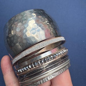 以下が含まれることがあります： ハンマー仕上げの幅広カフ、グリッター加工のバンド、滑らかなポリッシュ仕上げのバンド、ファセットカットビーズのブレスレット、細いワイヤー巻きのバンド、プレーンなバンド、幾何学模様が刻印された細いバンドが重なったシルバー調のメタルバングルセット。