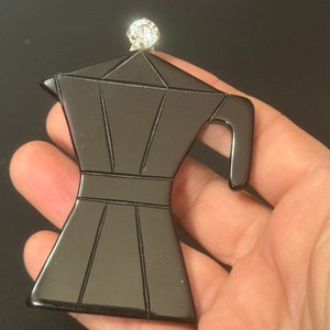 May include: A black resin brooch shaped like a coffee pot with a clear rhinestone on top.