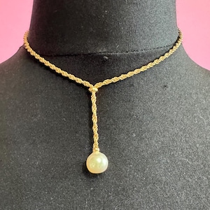 May include: A gold-colored necklace with a rope chain design. The necklace features a single, round, off-white pearl pendant. The necklace is displayed on a black mannequin against a pink background.