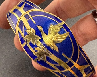 May include: A hinged bracelet with a vibrant blue enamel surface, accented with gold-colored metal. The design features a flying bird and floral motifs. The bracelet is held in a hand, showcasing its intricate details and craftsmanship.