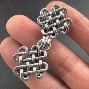 May include: A silver-toned metal clasp featuring a Celtic knot design. The clasp is composed of two intricately woven knot shapes connected by a small chain. The metal has a slightly aged appearance.