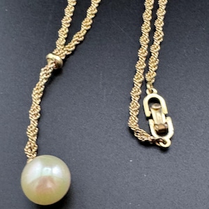 May include: A gold-toned necklace with a twisted rope chain design. The necklace features a large, round, off-white pearl pendant. The clasp is a simple, secure design. The necklace is displayed against a dark background.