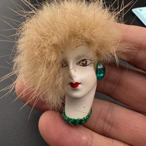 May include: A vintage brooch featuring a stylized face with a fur-like blonde hairpiece. The face has red lips, a green earring, and a green collar. The brooch is likely made of ceramic or porcelain.