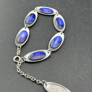 May include: Silver bracelet with oval links, each set with a vibrant blue gemstone. The bracelet has a chain and a decorative oval charm. The jewellery is shown against a dark backdrop.