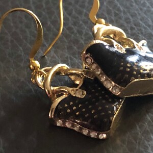 Puede incluir: Un par de pendientes de tono dorado con un dise&ntilde;o de bolso de mano de esmalte negro. Los bolsos de mano tienen un ribete de tono dorado y est&aacute;n acentuados con diamantes de imitaci&oacute;n transparentes.