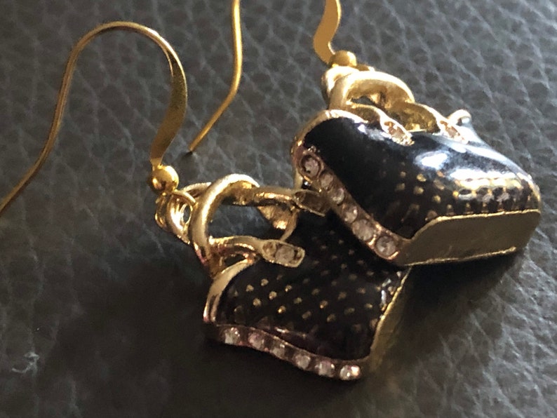 Puede incluir: Un par de pendientes de tono dorado con un dise&ntilde;o de bolso de mano de esmalte negro. Los bolsos de mano tienen un ribete de tono dorado y est&aacute;n acentuados con diamantes de imitaci&oacute;n transparentes.