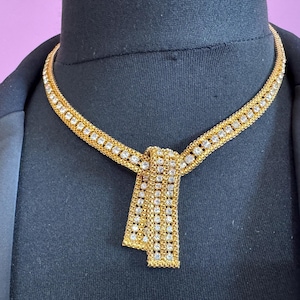 May include: A gold-toned necklace with a knot design, adorned with numerous clear rhinestones. The necklace is displayed on a black mannequin, showcasing its intricate details and sparkling appearance. The necklace is a statement piece.