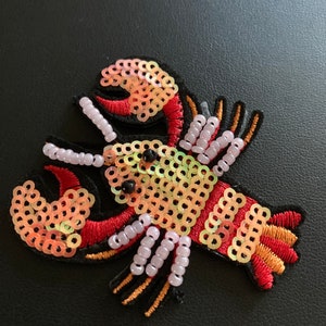 May include: An embroidered lobster patch in red, orange and yellow with iridescent sequins. The lobster is outlined in black thread and has a white bead border.
