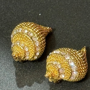 May include: A pair of gold-coloured seashell-shaped clip-on earrings. The earrings have a textured surface with small bumps and a spiral of clear rhinestones. The earrings have a vintage aesthetic.