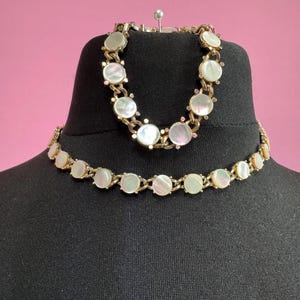 May include: A set of two gold tone necklaces with white mother of pearl round accents. The necklaces are on a black mannequin.