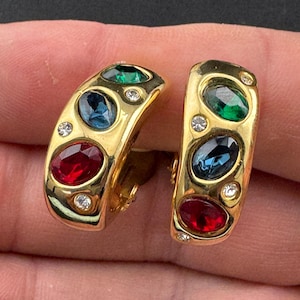 May include: Gold-coloured clip-on earrings featuring red, blue, and green oval gemstones. Small clear crystals are set around the gemstones. The earrings have a curved, rectangular shape.