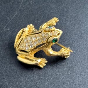 May include: A gold-coloured frog brooch with clear and green gemstones. The frog is detailed with clear rhinestones on its body and green stones for the eyes. The brooch has a pin closure on the back.