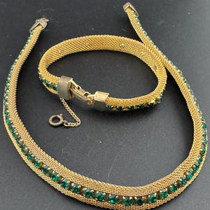 May include: A gold-toned mesh necklace and bracelet set. The jewelry is adorned with rows of small, round, emerald-green gemstones. The necklace has a small clasp and a delicate chain. The bracelet has a matching clasp.