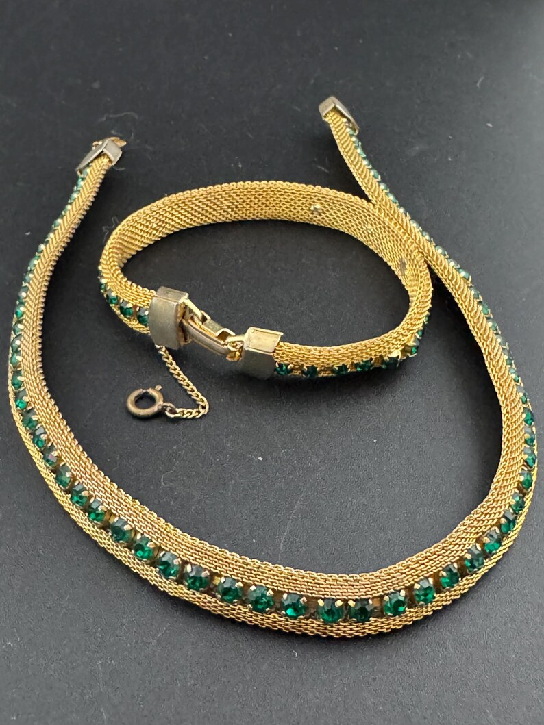 May include: A gold-toned mesh necklace and bracelet set. The jewelry is adorned with rows of small, round, emerald-green gemstones. The necklace has a small clasp and a delicate chain. The bracelet has a matching clasp.