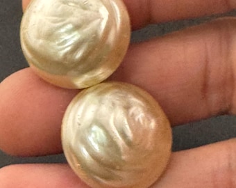 May include: Two large, round, pearlescent buttons with a swirling, textured surface. The buttons have a creamy, off-white color and a subtle sheen, suggesting a vintage or antique style. They are held in a hand, ready for sewing or crafting.