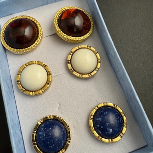 May include: Six vintage button earrings in a light blue box. The earrings have gold-colored metal frames. Two are dark brown, two are white, and two are dark blue with white speckles. The earrings are round and have a domed center.
