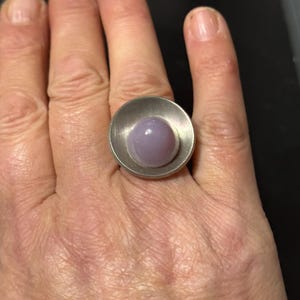 May include: A silver ring featuring a large, round, pale purple stone set in a silver bezel. The ring is worn on a person's finger.