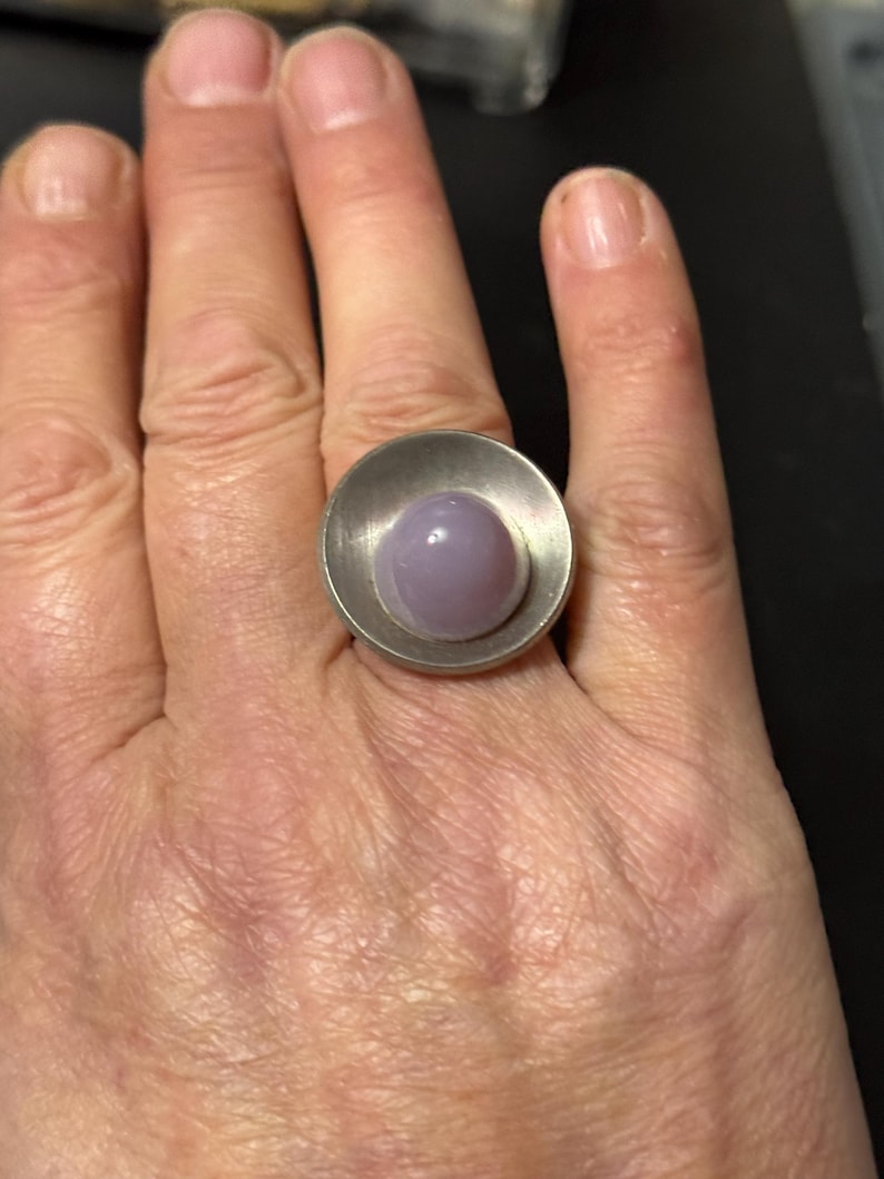 May include: A silver ring featuring a large, round, pale purple stone set in a silver bezel. The ring is worn on a person's finger.