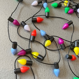 May include: A string of colorful Christmas lights with various colors including green, pink, yellow, red, white, and blue. The lights are strung on a black wire and are arranged on a gray surface. The lights are shaped like teardrops.