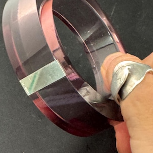 Könnte beinhalten: Zwei durchscheinende, dunkelviolette Armreifen, einer in dem anderen, sind abgebildet. Ein silbernes Metallschild ist an einem Armreif befestigt. Die Armreifen befinden sich auf einer dunklen Oberfläche. Das Bild ist eine Nahaufnahme der Armreifen.