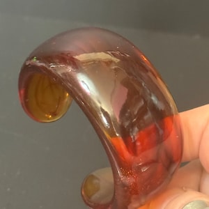 May include: A thick, amber-coloured glass bangle bracelet. The bracelet is smooth and shiny, with a slight iridescent quality.