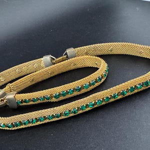 May include: Gold-toned mesh necklace with a row of emerald green rhinestones. The necklace has a clasp closure and is displayed on a dark surface. The jewelry piece is a vintage accessory.