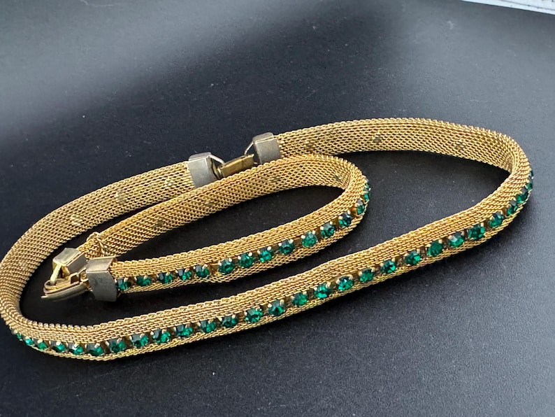 May include: Gold-toned mesh necklace with a row of emerald green rhinestones. The necklace has a clasp closure and is displayed on a dark surface. The jewelry piece is a vintage accessory.