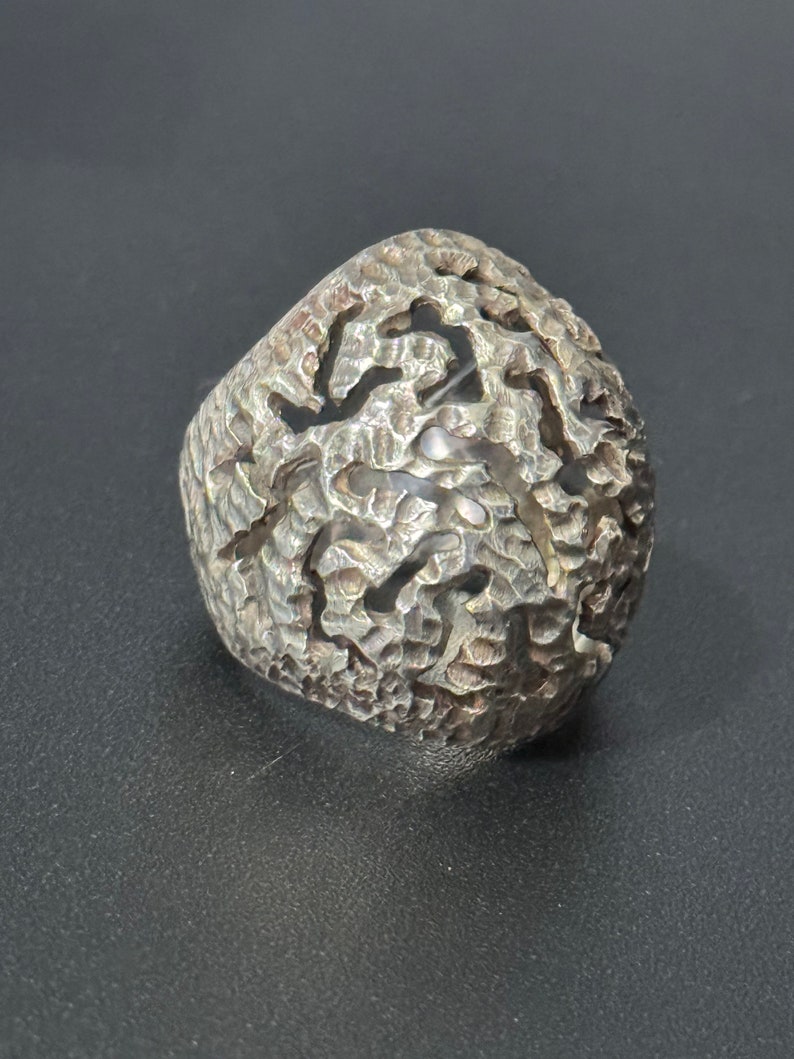 May include: A silver ring with a textured, abstract design. The ring is wide and has a unique, organic shape.