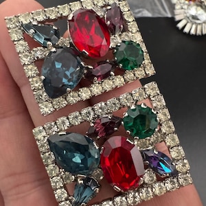 May include: Two square-shaped vintage rhinestone brooches. Each brooch features a silver-tone metal frame encasing a cluster of colorful, faceted gemstones in red, blue, green, and purple hues. The brooches are set with clear rhinestones around the edges.