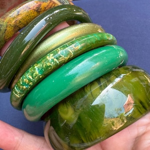 May include: A selection of green bangles in diverse shades, from olive to vibrant green, featuring unique patterns and textures. Some bangles have a glossy finish, while others appear matte. The bangles are stacked, displaying their varied designs.