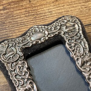 Puede incluir: Un marco de fotos plateado con un dise&ntilde;o ornamentado. El marco est&aacute; hecho de metal y tiene un respaldo de terciopelo negro.