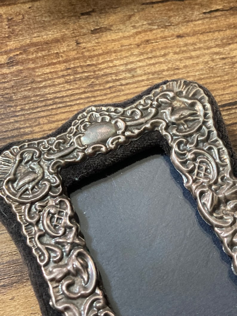 Puede incluir: Un marco de fotos plateado con un dise&ntilde;o ornamentado. El marco est&aacute; hecho de metal y tiene un respaldo de terciopelo negro.