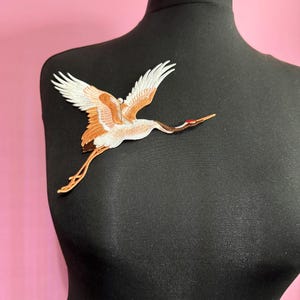 May include: An embroidered crane applique patch in brown and white. The crane is in flight with its wings spread and its head turned to the side. The patch is on a black fabric background.