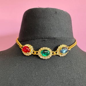 May include: A gold necklace featuring three oval gemstones in red, green, and blue. Each gem is encircled by a halo of small, clear crystals. The necklace is presented on a black mannequin head, set against a pink backdrop.