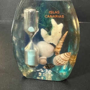 May include: A clear glass paperweight containing a small hourglass, seashells, and a piece of coral. The text "ISLAS CANARIAS" is printed on the top of the paperweight.