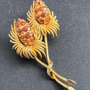 May include: A gold-toned brooch in the shape of two stylized flowers. Each flower has a central cluster of orange-brown rhinestones surrounded by detailed, radiating petals. The stems are intertwined, creating an elegant, nature-inspired design.