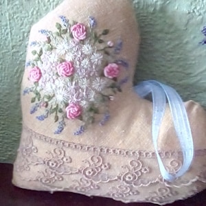 Op de afbeelding: Een beige hartvormig kussen van stof met een geborduurd bloemenmotief met roze en witte bloemen en blauwe bladeren. Het kussen heeft een blauw satijnen lint eromheen gebonden en een kantversiering aan de rand.