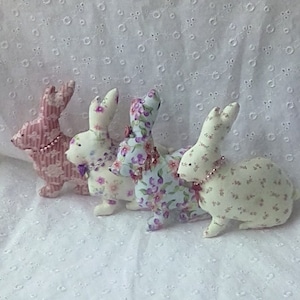 Puede incluir: Cuatro conejos de tela con estampados florales. Los conejos presentan estampados florales rosas, morados y azules. Cada conejo tiene un collar de cuentas y un pequeño lazo. El fondo es una tela blanca texturizada. Los conejos son de tela.