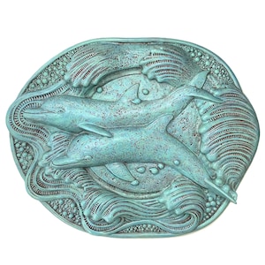 Puede incluir: Una placa de forma ovalada con una escultura en relieve de dos delfines nadando entre olas estilizadas. La pieza presenta un acabado desgastado en color verde azulado con acentos oscuros en las áreas hundidas, resaltando la textura.