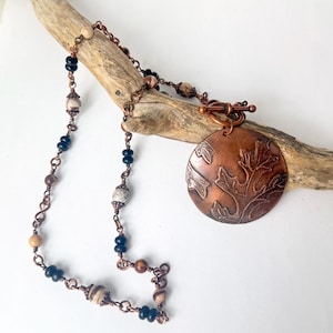 May include: A necklace featuring a round copper pendant with an embossed leaf pattern, attached to a copper chain. The chain is accented with alternating black beads and round, multi-colored stone beads, finished with a copper toggle clasp.