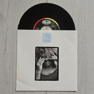 Pode incluir: Um disco de vinil de 7 polegadas numa capa de papel branco com o texto TINA TURNER PRIVATE DANCER. A capa apresenta uma fotografia a preto e branco de uma pessoa a ajustar os óculos de sol. O rótulo do disco é preto e colorido.