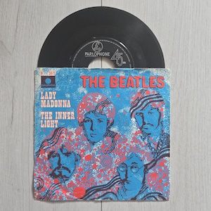 Pode incluir: Um disco de vinil de 7 polegadas dos Beatles com as músicas Lady Madonna e The Inner Light. A capa de papel quadrada exibe uma ilustração gráfica azul e vermelha dos quatro membros da banda com o texto The Beatles, Lady Madonna e The Inner Light impresso na frente. O disco de vinil preto é parcialmente visível, saindo pela parte superior da capa.