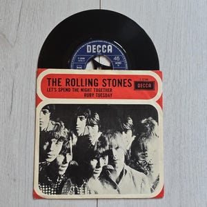 Pode incluir: Um disco de vinil preto de 45 RPM dos Rolling Stones numa capa de papel. A capa apresenta uma fotografia a preto e branco dos membros da banda e texto vermelho que diz THE ROLLING STONES, LET'S SPEND THE NIGHT TOGETHER e RUBY TUESDAY.
