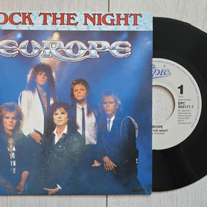 Pode incluir: Um disco de vinil de 7 polegadas da música Rock the Night da banda Europe. A capa de papel quadrada apresenta uma fotografia em tons de azul dos cinco membros da banda com cabelos longos, e o disco preto tem um rótulo central branco.