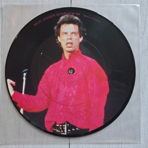 Pode incluir: Um disco de vinil ilustrado circular com uma fotografia de Mick Jagger vestindo uma camisa vermelha brilhante. O texto "MICK JAGGER-INTERVIEW 89" e "Made in England" está impresso na borda superior. O disco está em uma capa de plástico.