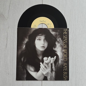 Pode incluir: Um disco de vinil de 7 polegadas de The Sensual World de Kate Bush. A capa de papel quadrada apresenta um retrato em preto e branco de uma pessoa com cabelos longos e escuros segurando um pequeno objeto. O texto diz The Sensual World Kate Bush em dourado.