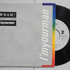 Pode incluir: Um disco de vinil de 7 polegadas numa capa de papel cinzenta com o texto WHAM! e I'm your man. A capa tem uma faixa vertical com as cores do arco-íris no lado direito. O disco preto é parcialmente visível, mostrando um rótulo branco central.