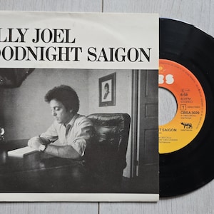 Pode incluir: Um disco de vinil de 7 polegadas de Goodnight Saigon de Billy Joel. A capa de papel branco apresenta uma fotografia a preto e branco de uma pessoa sentada a uma secretária. O disco tem uma superfície preta com sulcos e um rótulo central laranja e amarelo.