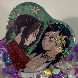 May include: A heart-shaped button with a holographic finish features two women with long dark hair. The woman on the left is wearing a red shirt with white hearts and the woman on the right is wearing a white shirt with a yellow flower in her hair. The background is green with white flowers.
